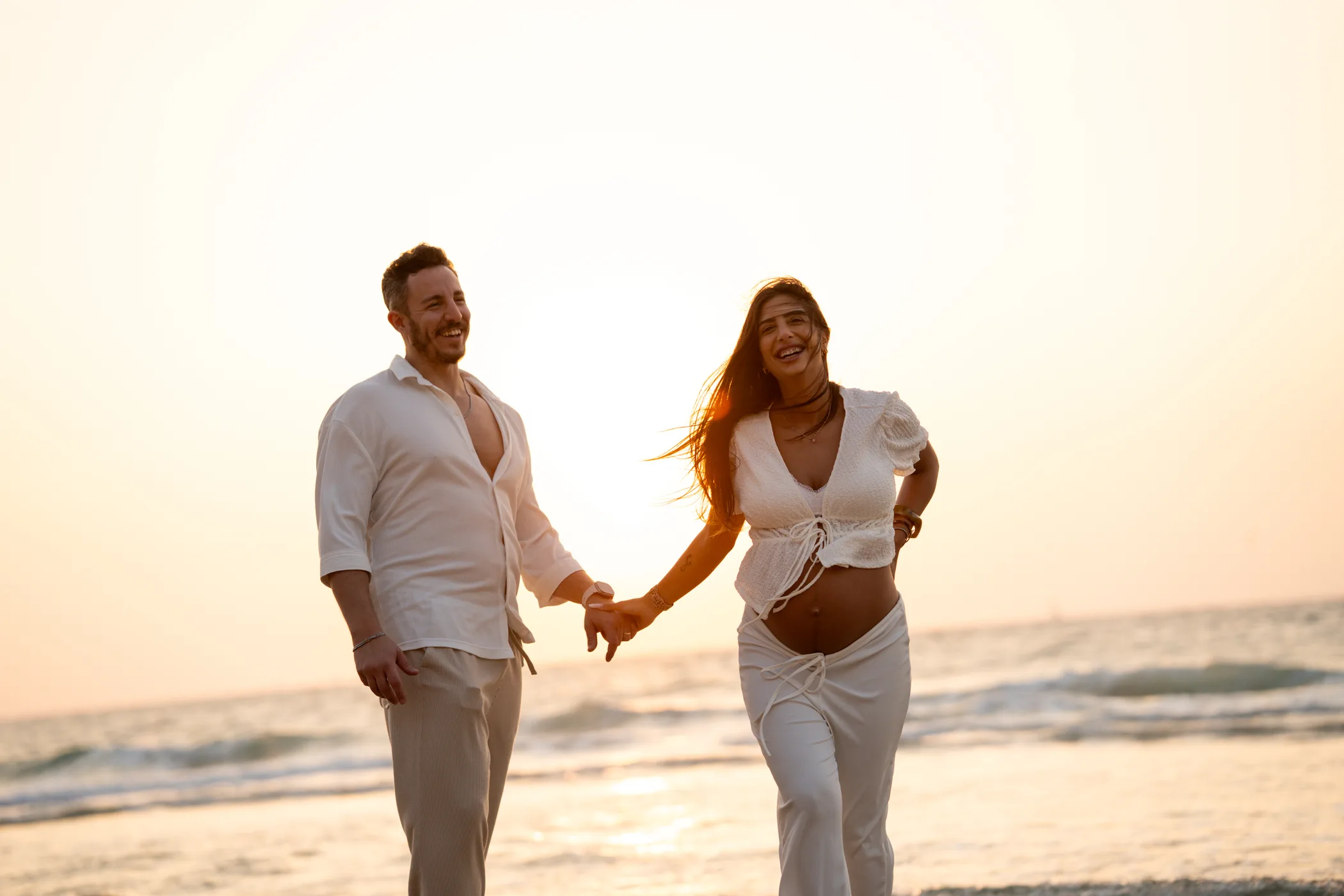 An expectant couple walking along the shore at sunset, laughing